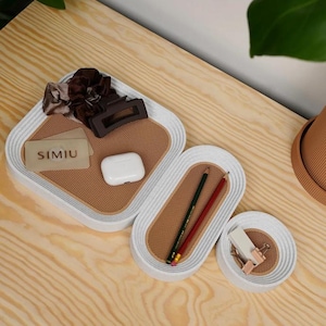 May include: A set of four white and tan decorative trays of varying sizes. The trays have a textured tan interior and a white, ridged border. Items inside include hair accessories, pencils, and a business card with the text "SIMIU".