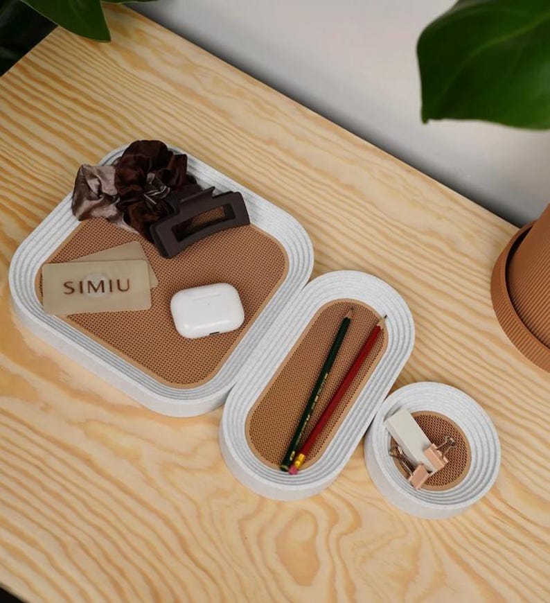 May include: A set of four white and tan decorative trays of varying sizes. The trays have a textured tan interior and a white, ridged border. Items inside include hair accessories, pencils, and a business card with the text "SIMIU".