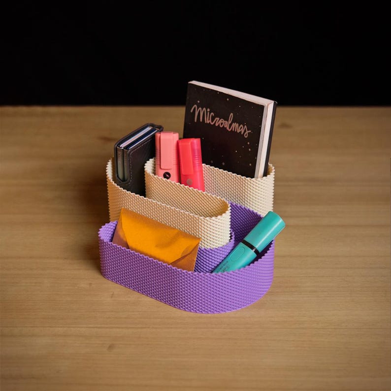 May include: A desk organiser with a wavy, textured structure featuring a cream-coloured upper section and a purple lower section. It holds a black notebook with the text "Microalmas" on the cover, a black case, two pink markers, a gold envelope, and a teal pen.