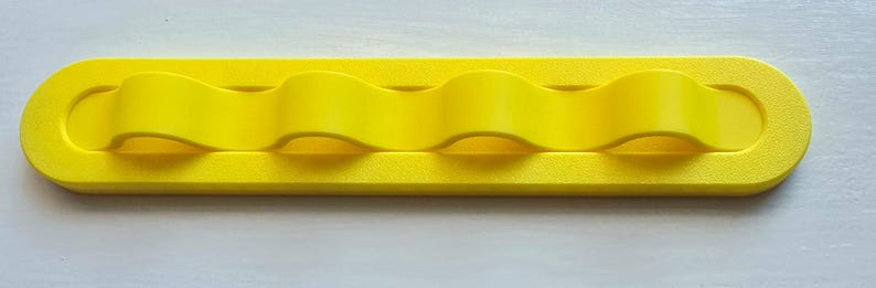 May include: A bright yellow, elongated, rectangular wall-mounted organizer featuring four distinct, wave-shaped loops integrated into the center of the frame. The object has a smooth, matte finish and rounded ends, designed for holding items securely.