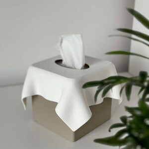 May include: A square tissue box with a white, draped top and a beige base. White tissues are pulled from the center opening. The design is minimalist and modern, suitable for home decor.
