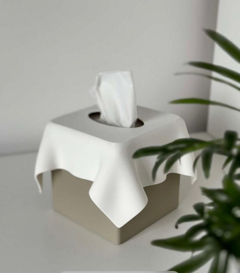 May include: A square tissue box with a white, draped top and a beige base. White tissues are pulled from the center opening. The design is minimalist and modern, suitable for home decor.