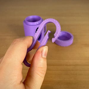 May include: A purple 3D-printed carabiner clip with a textured surface, held in front of a matching container and lid. The clip is open, showing its hook design. The container and lid have a similar textured pattern.