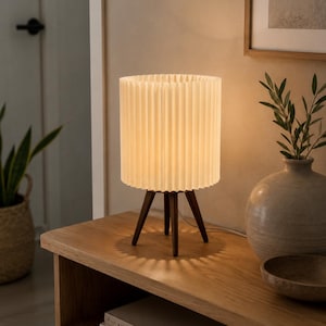 Puede incluir: Una l&aacute;mpara de mesa con una pantalla cil&iacute;ndrica blanca plisada, sostenida por tres patas de madera de color marr&oacute;n oscuro. La l&aacute;mpara emite una luz c&aacute;lida y suave, proyectando un patr&oacute;n de sombra en forma de estrella bajo su base.