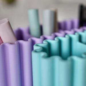 K&ouml;nnte beinhalten: Nahaufnahme eines mehrfarbigen Beh&auml;lters mit gewelltem Rand. Der Beh&auml;lter ist in Abschnitte aus Helllila, T&uuml;rkis und Hellblau unterteilt. Zylindrische Objekte in Rosa-, Hellblau- und Graut&ouml;nen sind im Inneren sichtbar. Der Beh&auml;lter scheint aus Silikon zu bestehen.