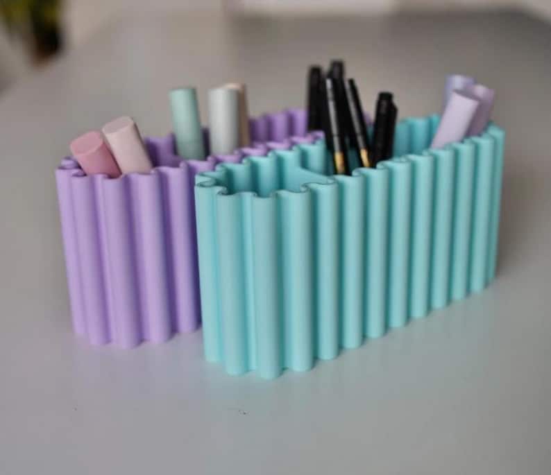 K&ouml;nnte beinhalten: Zwei pastellfarbene Beh&auml;lter mit gewelltem Rand. Einer ist lavendelfarben, der andere aquamarin. Die Beh&auml;lter enthalten verschiedene Make-up-Artikel, darunter Lippenstifte, Bleistifte und Stifte. Die Beh&auml;lter stehen auf einer hellgrauen Oberfl&auml;che.
