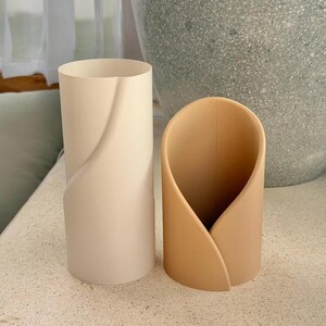 May include: Two cylindrical containers with a smooth, matte finish. The taller container is off-white and features a subtle diagonal seam. The shorter container is a warm tan color and has a curved, overlapping design that creates an open front.