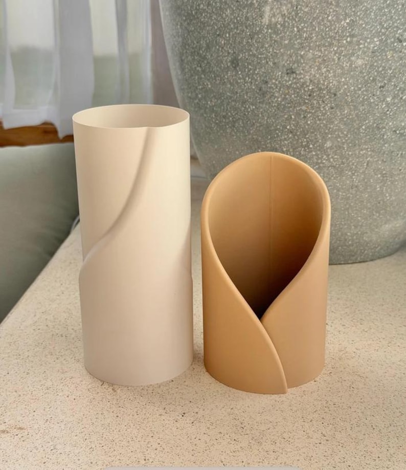 May include: Two cylindrical containers with a smooth, matte finish. The taller container is off-white and features a subtle diagonal seam. The shorter container is a warm tan color and has a curved, overlapping design that creates an open front.