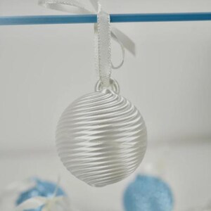 Pode incluir: Um ornamento esf&eacute;rico branco com um padr&atilde;o texturizado de nervuras em espiral. O ornamento possui um pequeno la&ccedil;o no topo, ao qual est&aacute; atada uma fita de cetim branca, permitindo que fique pendurado numa haste horizontal azul.