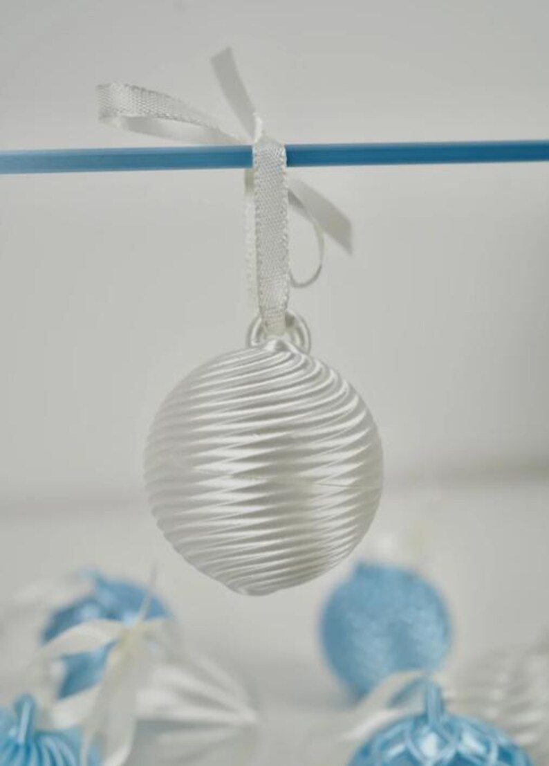 Pode incluir: Um ornamento esf&eacute;rico branco com um padr&atilde;o texturizado de nervuras em espiral. O ornamento possui um pequeno la&ccedil;o no topo, ao qual est&aacute; atada uma fita de cetim branca, permitindo que fique pendurado numa haste horizontal azul.