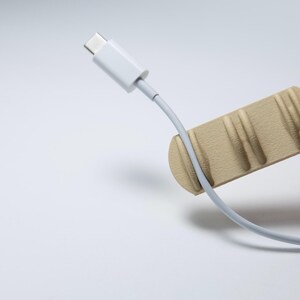 May include: A light beige cable organiser with a textured, ridged surface holds a white USB-C charging cable. The cable is positioned vertically, with the connector end pointing upwards, resting securely within one of the moulded grooves of the holder.