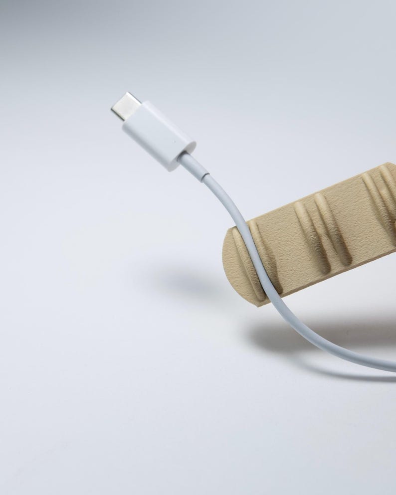 May include: A light beige cable organiser with a textured, ridged surface holds a white USB-C charging cable. The cable is positioned vertically, with the connector end pointing upwards, resting securely within one of the moulded grooves of the holder.
