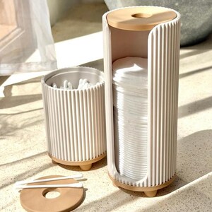 May include: A cylindrical white ribbed cotton pad dispenser with a tan base and top lid, standing next to a smaller matching cylindrical container filled with white cotton swabs. A separate tan lid with a center hole lies flat on the surface.