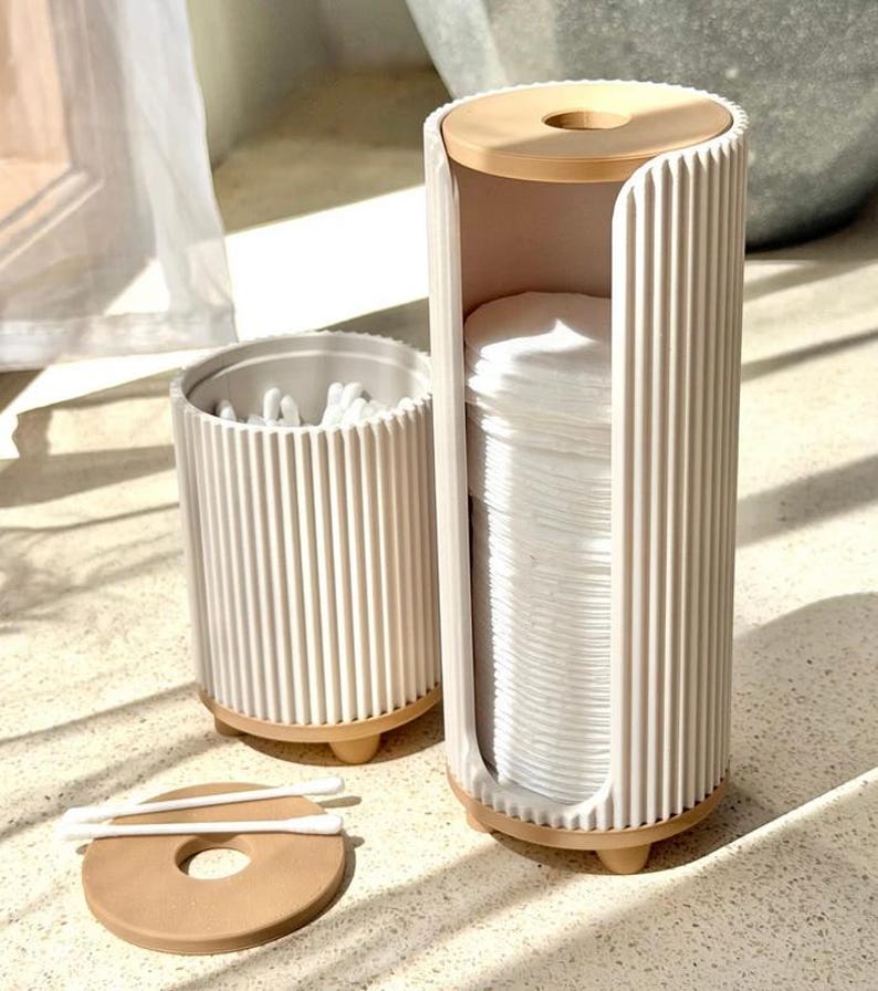 May include: A cylindrical white ribbed cotton pad dispenser with a tan base and top lid, standing next to a smaller matching cylindrical container filled with white cotton swabs. A separate tan lid with a center hole lies flat on the surface.