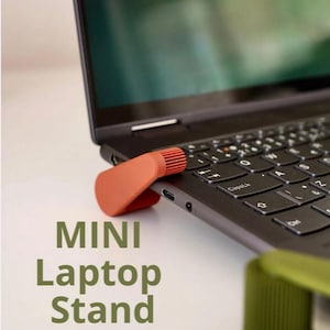 May include: An orange mini laptop stand is attached to the side of a black laptop. The stand has a curved design with a ribbed top. The words "MINI Laptop Stand" are printed in green below the laptop.