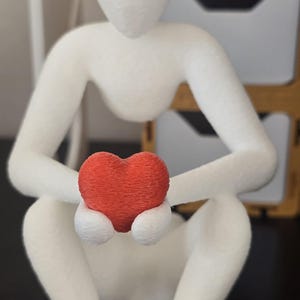 Puede incluir: Figura blanca de una persona sentada sosteniendo un coraz&oacute;n rojo. La figura est&aacute; hecha de un material texturizado, y el coraz&oacute;n es de un rojo vibrante. La pose sugiere contemplaci&oacute;n o cuidado. La figura mide unos 15 cm de altura.