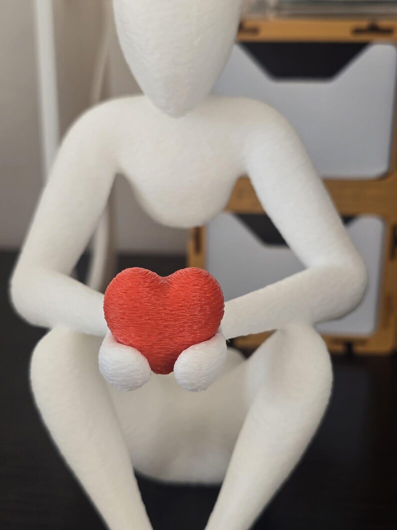 Puede incluir: Figura blanca de una persona sentada sosteniendo un coraz&oacute;n rojo. La figura est&aacute; hecha de un material texturizado, y el coraz&oacute;n es de un rojo vibrante. La pose sugiere contemplaci&oacute;n o cuidado. La figura mide unos 15 cm de altura.