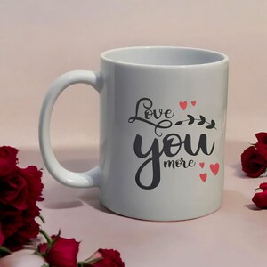 May include: A white ceramic mug with a curved handle. The mug features the phrase "Love you more" in black script with red heart accents. The mug is surrounded by red roses.