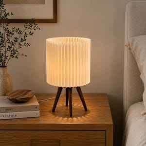 Puede incluir: Una l&aacute;mpara de mesa con una pantalla cil&iacute;ndrica plisada de color blanco roto, sostenida por tres patas de madera de color marr&oacute;n oscuro. La l&aacute;mpara emite una luz c&aacute;lida y suave, proyectando una sombra en forma de estrella sobre la superficie.