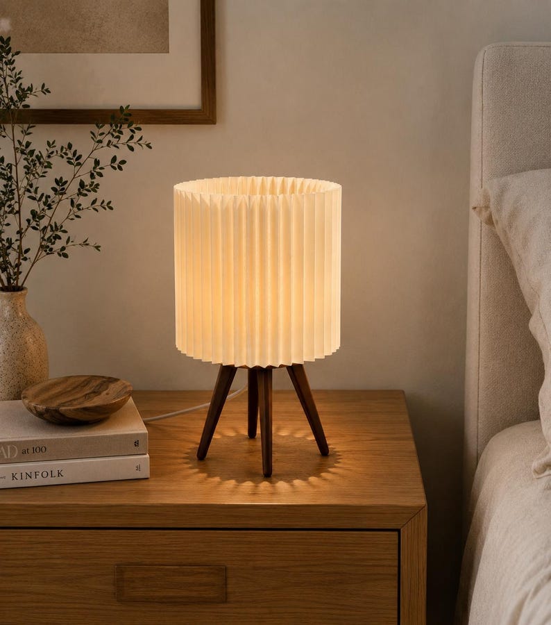 Puede incluir: Una l&aacute;mpara de mesa con una pantalla cil&iacute;ndrica plisada de color blanco roto, sostenida por tres patas de madera de color marr&oacute;n oscuro. La l&aacute;mpara emite una luz c&aacute;lida y suave, proyectando una sombra en forma de estrella sobre la superficie.