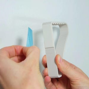 May include: A light gray tape dispenser with a curved design, held near a piece of blue painter's tape. The dispenser has a series of parallel lines on one side and a rectangular section with a logo. The background is a plain white wall.