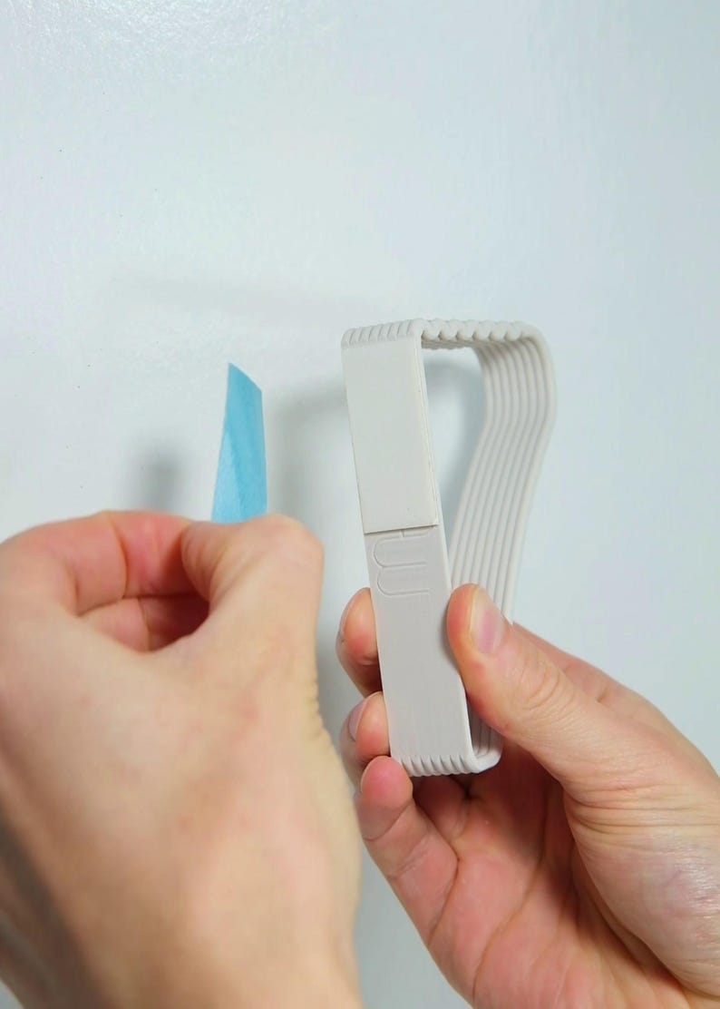 May include: A light gray tape dispenser with a curved design, held near a piece of blue painter's tape. The dispenser has a series of parallel lines on one side and a rectangular section with a logo. The background is a plain white wall.