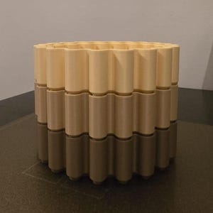 May include: A three-tiered, cylindrical decorative container. The top tier is a light beige color, the middle tier is a medium beige, and the bottom tier is a dark beige. The container has a wavy, repeating pattern around its circumference.