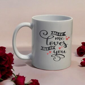 May include: A white ceramic mug with a curved handle. The mug features the text "All of me loves all of you" in black script with small red heart accents. The mug is surrounded by red roses.