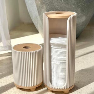 May include: Two cylindrical white ribbed storage containers with light tan circular lids and small feet. The taller container features a vertical opening displaying a stack of white cotton pads, while the shorter container is closed.
