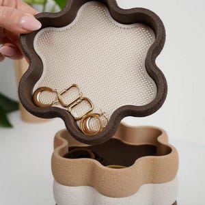Puede incluir: Una joyero de varios niveles con un dise&ntilde;o en forma de flor. La capa superior, abierta, revela una superficie de malla beige y pendientes de aro dorados. La caja est&aacute; compuesta por capas marrones, beige y blancas, creando una apariencia texturizada y en capas.