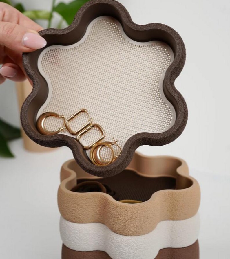 Puede incluir: Una joyero de varios niveles con un dise&ntilde;o en forma de flor. La capa superior, abierta, revela una superficie de malla beige y pendientes de aro dorados. La caja est&aacute; compuesta por capas marrones, beige y blancas, creando una apariencia texturizada y en capas.