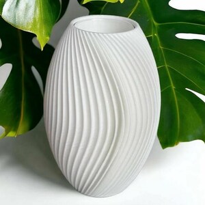 May include: A white, oval-shaped vase with a textured, ridged surface. The vase has a wide opening and a smooth interior. The background features large green leaves. The vase is a decorative item.