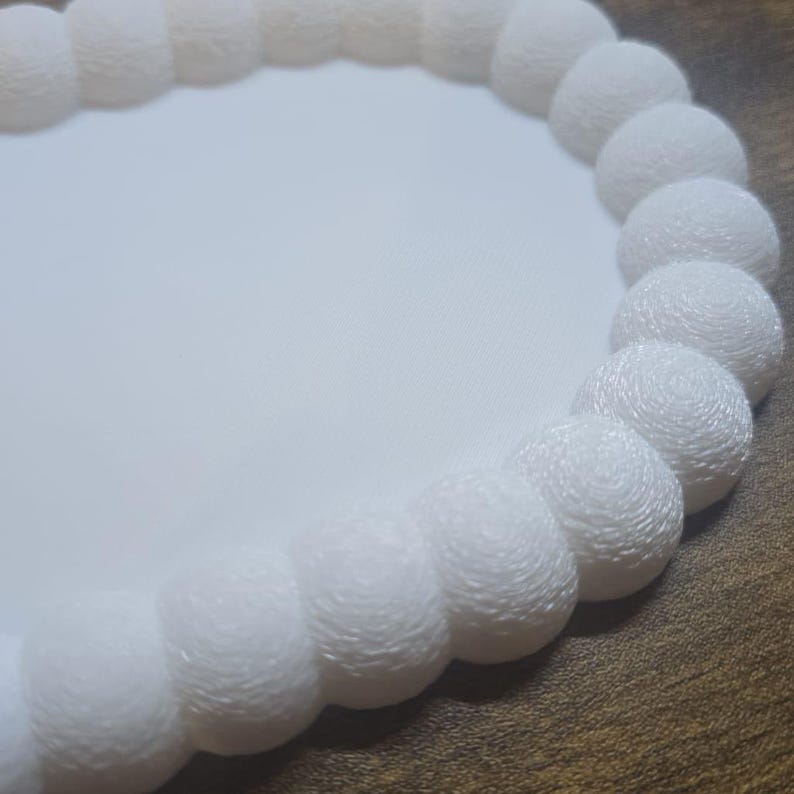 May include: A white, round placemat with a textured, beaded edge. The placemat has a smooth, plain center and a decorative border of evenly spaced, spherical beads. The design is simple and elegant.