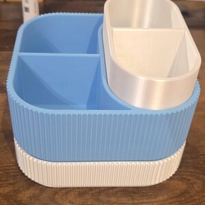 May include: A two-tone desk organizer with a light blue and white color scheme. The organizer has multiple compartments, including a larger section with dividers and a smaller, oval-shaped section. The exterior features a ribbed design.