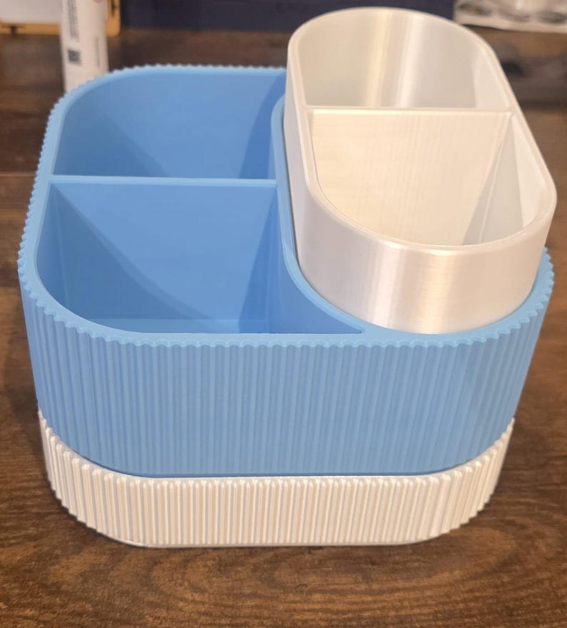 May include: A two-tone desk organizer with a light blue and white color scheme. The organizer has multiple compartments, including a larger section with dividers and a smaller, oval-shaped section. The exterior features a ribbed design.