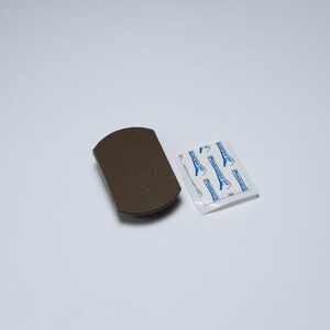 May include: A dark brown, rectangular adhesive mounting hook base with rounded edges, accompanied by a small, square white adhesive strip backing featuring blue text that reads Powerstrips in a repeating pattern across the surface.