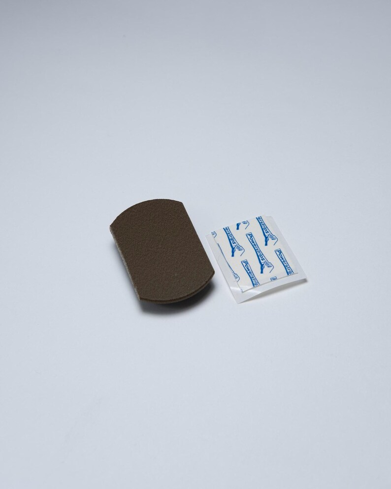 May include: A dark brown, rectangular adhesive mounting hook base with rounded edges, accompanied by a small, square white adhesive strip backing featuring blue text that reads Powerstrips in a repeating pattern across the surface.