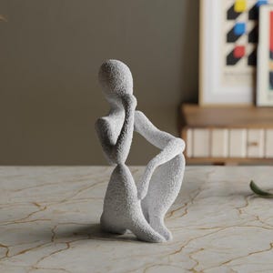 Puede incluir: Una escultura texturizada, de color gris claro, de una persona en una pose sentada y contemplativa. La figura est&aacute; arrodillada con un brazo apoyado en una rodilla doblada y la otra mano tocando la cara. La escultura est&aacute; sobre una superficie similar al m&aacute;rmol.