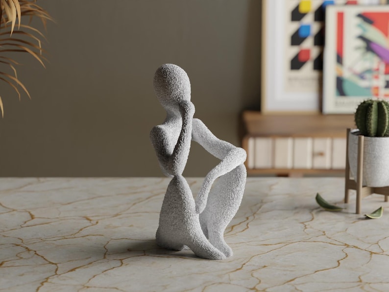 Puede incluir: Una escultura texturizada, de color gris claro, de una persona en una pose sentada y contemplativa. La figura est&aacute; arrodillada con un brazo apoyado en una rodilla doblada y la otra mano tocando la cara. La escultura est&aacute; sobre una superficie similar al m&aacute;rmol.