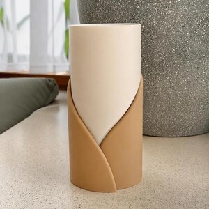 May include: A cylindrical vase featuring a smooth, off-white inner vessel partially encased by a matte, tan-colored outer sleeve that wraps around the base in a curved, overlapping design, creating a layered appearance with a clean, vertical profile.