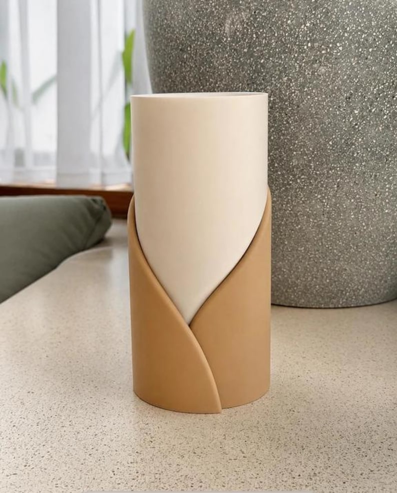 May include: A cylindrical vase featuring a smooth, off-white inner vessel partially encased by a matte, tan-colored outer sleeve that wraps around the base in a curved, overlapping design, creating a layered appearance with a clean, vertical profile.