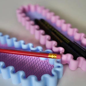 May include: Two pastel-colored pencil cases with a wavy edge design. One case is blue and the other is pink. A red pencil with a gold ferrule and eraser is in the blue case. Black pens are in the pink case.