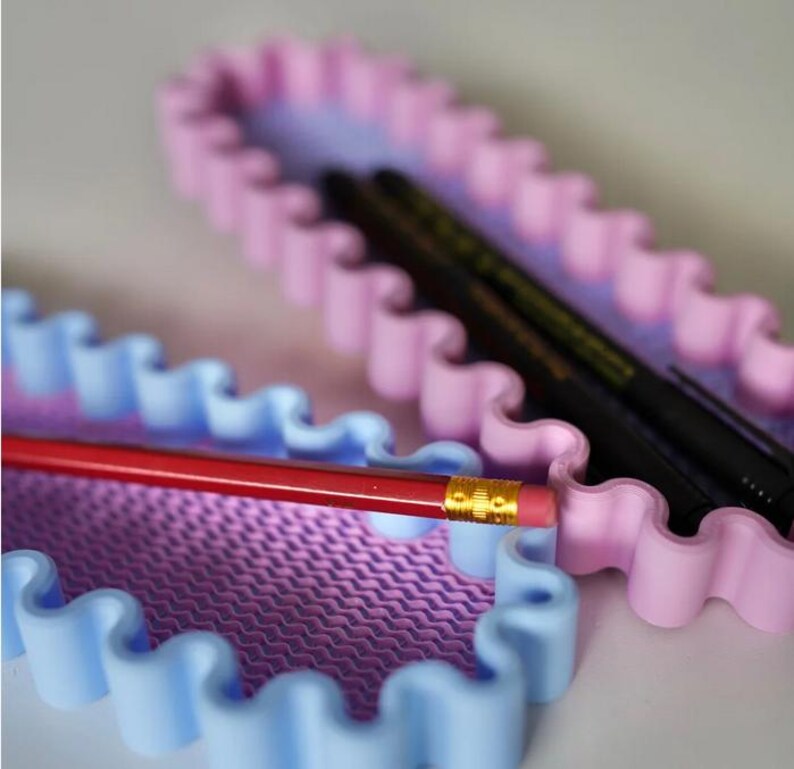 May include: Two pastel-colored pencil cases with a wavy edge design. One case is blue and the other is pink. A red pencil with a gold ferrule and eraser is in the blue case. Black pens are in the pink case.
