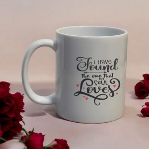 May include: White ceramic mug with a large handle. The mug features the text "I have found the one that my soul loves" in a black, cursive font with small red heart accents. The mug is surrounded by red roses.