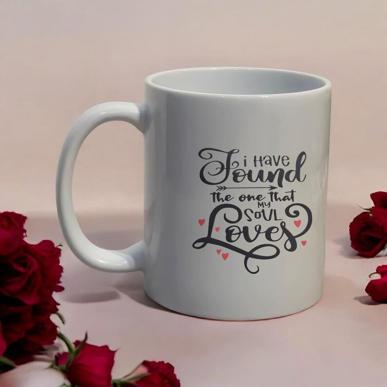 May include: White ceramic mug with a large handle. The mug features the text "I have found the one that my soul loves" in a black, cursive font with small red heart accents. The mug is surrounded by red roses.