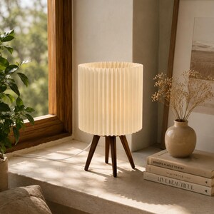 Puede incluir: Una l&aacute;mpara de mesa con una pantalla cil&iacute;ndrica plisada de color crema y tres patas de madera marr&oacute;n oscuro. La l&aacute;mpara est&aacute; encendida, emitiendo un brillo suave, y cuenta con un cable de alimentaci&oacute;n blanco que sale de la base.