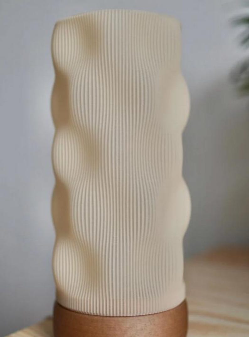 May include: A beige, textured vase with a ribbed design. The vase has a rounded, organic shape with a wooden base. The vertical lines create a visually interesting pattern. The neutral color palette makes it suitable for various decor styles.
