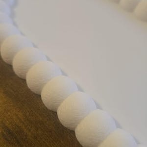 May include: A white, oval-shaped decorative tray with a textured, beaded border. The tray's interior is smooth and white, contrasting with the detailed, spherical border. The tray sits on a brown wooden surface.