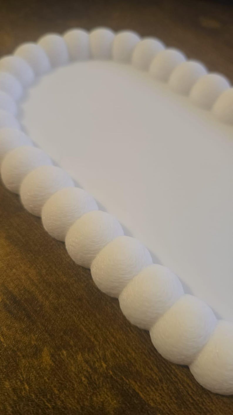 May include: A white, oval-shaped decorative tray with a textured, beaded border. The tray's interior is smooth and white, contrasting with the detailed, spherical border. The tray sits on a brown wooden surface.
