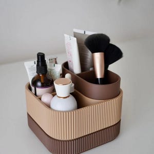 May include: A two-tiered, brown and beige cosmetic organizer with vertical ribbed detailing. The organizer holds various beauty products, including brushes, tubes, and bottles. The top tier is a lighter beige, while the bottom tier is a darker brown.